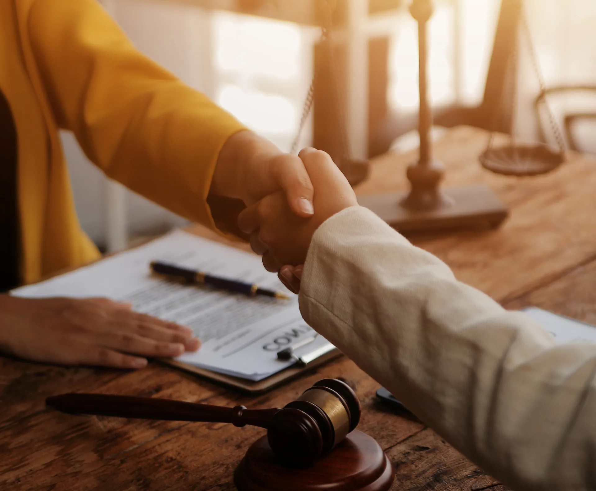 Attorney and client shaking hands over a signed agreement with a gavel and scales of justice on the desk